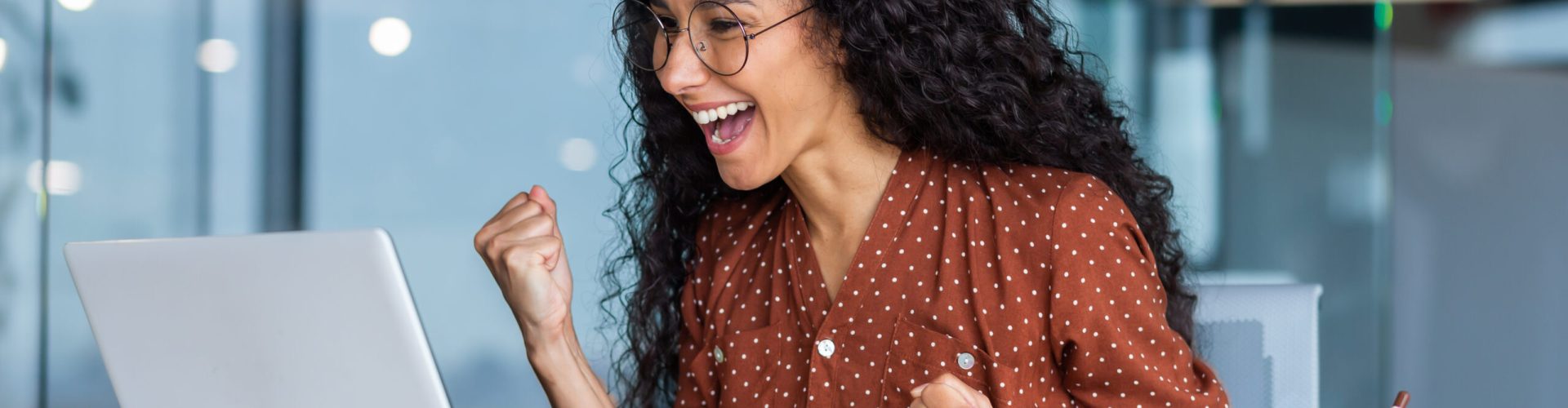 Happy beautiful young Latin American woman sitting in the office in front of the laptop monitor, happy about the win, good news, victory. He smiles, shows a yes gesture with his hands.