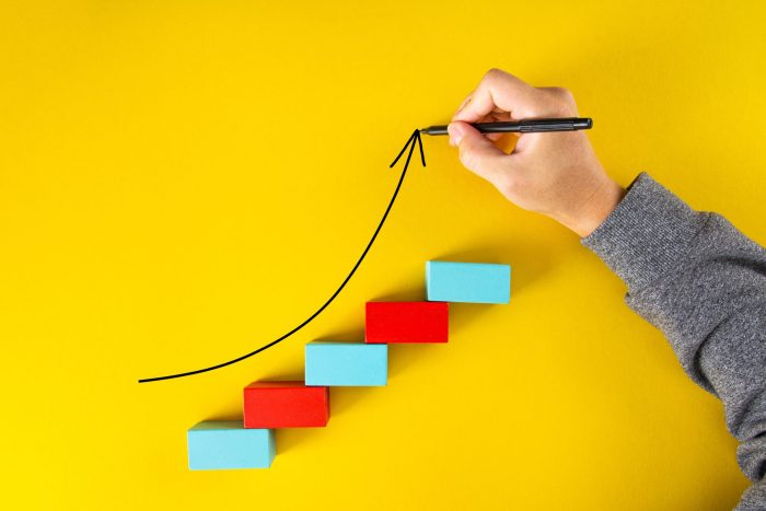 Male hand drawing an upward pointing arrow on top of growing graph made of wooden blocks over yellow background